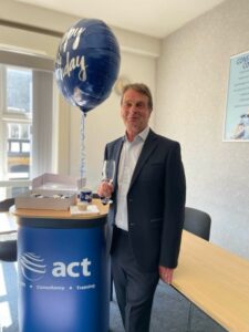 ACT Managing Director and founder Ian Coombes holds a glass of champage next to a blue foil balloon that says Happy Birthday